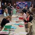 Several preteen and teenage girls creating Christmas cards at a long table in the church hall. A few adults are standing or sitting towards the back of the photo.