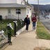 A woman and a man standing on the sideward next to the wrought iron fence that surrounds the backyard of the rectory. They have just finished putting large white bows every few feet along the length of the fence.