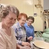Three women standing in a line that runs from the lower-left corner to roughly 2/3 of the way to the upper-right corner. They're standing at a counter in the kitchen of the church hall and there are various bowls and a large pastel-green dough mixer in view in front of them, and running along the lower-right corner of the frame. The women are smiling and mostly looking away from the camera.