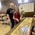 Four workers are standing in front of a table that runs from the foreground to mid-way up the left-side of the frame. There are three Silpat mats on the table and, on top of the mats, there are several pieces of dough that are rolled into long strands that look almost like snakes. Some are very long, extending across more that one mat, and short pieces that fit within the length of their mat. The workers are looking at the camera and smiling.