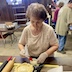 A woman is standing at a table, basting a paska bread with butter using a brush with a translucent white handle and red brush tendrils (sorry, I'm not sure what to call them, but I mean the actual brush part). She is facing the camera but is looking at her work. Two rolling pins are on the table to her right; one has a light brown roller and handle, while the other has a black roller body and a red handle. Only one handle is visible for each rolling pin.