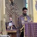 Fr. Aleksey appears in the right side of the frame and is reading from the bible that is on the lecturn in front of him. A white-haired priest can be seen standing behind the altar table in the lower left corner of the photo. His hands are clasped in front of him, and he's holding a white piece of paper. Three candles are on the altar table in front of him.