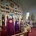 Six priests in purple vestments are standing in a straight line facing the iconostasis. They are standing between the center icon table and the two steps leading up to the walkway that runs the length of the iconostasis. The photo was taken to the left, and behind, the priest on the far left.