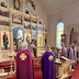 Five priests in purple vestments are standing in a straight line facing the iconostasis. They are standing between the center icon table and the two steps leading up to the walkway that runs the length of the iconostasis and facing Fr. Alexsey, who is standing in front of the Royal Doors while facing the priests and the congregation. The photo was taken to the left, and behind, the priest on the far left.