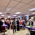 Parishioners and visitors standing for a prayer before the lenten meal begins. Three long tables with purple tablecloths run perpendicular to the lower right corner of the photo, and people are standing on either side of the tables, facing towards someone off-camera and left-of-frame.