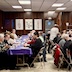 Parishioners and guests seated at tables in the church hall for a lenten meal. On the left two-thirds of the picture, a long table runs perpendicular from the bottom of the photo towards the back. Part of a second table is visible in the right third. The walls having brown paneling with some mostly white informational posters hanging on the back wall.