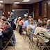 Parishioners and guests seated at tables in the church hall for a lenten meal. A long table runs from the lower-right corner to the center of the photo, and part of a second table is visible in the lower left corner. The walls having brown paneling, and there's a blackboard on the back wall. There's also a person to the left of the blackboard, who is walking toward the left side of the photo.