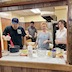 View of four people standing behind the counter in the kitchen of the church hall, posing for the camera. There are various ingredients and baking tools on the counter. The man on the far left is wearing a dark shirt and baseball cap, and he's stirring something in a black pot, using a large red, plastic spoon. Father Aleksey appears next but is standing further back in the kitchen. In the right half of the frame, two women are standing behind the same counter while smiling and posing for the camera. Everyone is wearing plastic food-prep gloves.