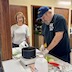 A man in a dark blue shirt and maching baseball cap is cutting up butter to put into a pot that rests in the center of the frame. A women with a white-and-gray-striped shirt is standing behind and partly to the left of the pot, around the corner from the man. She's wearing glasses and is smiling for the camera. An open half-gallon plastic container of milk appears in the center foreground, although the container is empty enough that no milk is visible.