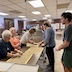 Four women are standing at the table mentioned in the previous photo. Three are standing on the left side, and one is opposite of them, standing on the right. They are making long dough braids, although there's a small dough cross in the picture too. There's also a young man standing along the right edge of the photo, and he's wearing a dark blue t-shirt with dark green shorts, along with clear, food prep gloves.