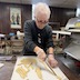 A woman in a black, long-sleeved shirt, with the sleeves pushed up, is standing behind a brown table with a white slicon mat on it. There are several pieces of dough on the mat, and they're cut into roughly 6-inch to 12-inch length, so of which are rolled. There's also a long, roughly two-foot rolled piece that runs from the bottom edge of the photo, and she's cutting it length-wise.
cutting long lengths of 