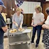 A woman is standing behind a three-level, metal serving cart and is cutting a finished paska bread for the baking crew to share. There are two paper plates on the top level of the cart. The one on the left has two quarter-loaves, and on the plate on the right, the woman is cutting another quarter of the bread into slices. Two women are visible on the right side of the frame as they look on, and a man partially appears on the left side, as he watches the woman cutting the bread. The bottom two levels of the serving cart are empty.