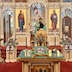 Aleksey is standing in front of the Royal Doors, facing the congregation, and with an altar server standing in front of him behind the center table. The alter server's back faces the camera, and he's wearing a gold vestment. The center table is covered in a green cloth. On the table, there's an icon that is now surrounded by a green cloth that serves as a frame. The table also holds two tall white candles in gold candle holders, two vigil lights, a gold cross, and some pussy willows. There are also two icon tables on either edge of the frame.