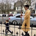 An altar server in a gold vestment is carrying a large, wooden cross as he walks to the left, outside of the church. There's a young boy walking ahead of him and holding a small, wooden cross. There's a black, wrought-iron fence in the foreground, and it's very ornate. In the background, a neighborhood house and several cars are visible.