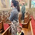 A woman and a young boy standing in a the last pew as viewed from the left side of the pew. The woman is facing towards the left side of the frame, looking towards the out-of-view front of the church, and the boy is smiling and looking at the camera. A man can be seen standing in the pew across the aisle, in the same row. A few pews can be seen in front of the people, and there's an off-white railing partially shown on the right side of the frame.