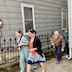 Part of the procession line outside of the church, where four people are visible, walking from the right side of the frame to left. Two woman are walking together and appear just to the left of the center of the photo. The woman on the left has her right arm in a sling, and the woman to her left (towards the center) is holding onto her arm. The second woman is possibly the mother of the first woman, who is wearing a teal jumpsuit with a pattern of white and black dots. The second woman is weating an orangey dress with blue, white, and teal flowers, and a deep blue bottom edge. She's also wearing a royal blue sweater. They're both carring pussy willows. The man behind them is wearing a medium brown shirt and dark blue pants. He's carrying and looking down at where he's stepping. A man behind him is wearing a green shirt and blue pants and is only partially visible.