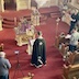 View from the choir loft, with Fr. Aleksey wearing a black vestment and standing in front of a lecturn while facing the iconostas. His lecturn sits in front of the center table, both of which are covered in purple cloths. There's a large candle holder, with at least a dozen candles on it between the lecturn and table. The carpeting is red, and a few parishioners in pews are visible on the left and right edges of the frame. This photo was taken from a position located above the left-hand-side pews, almost looking over Fr. Aleksey's left shoulder.
