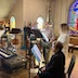 A view of part of the choir in the choir loft. A woman in a black coat is shown in profile, and she's seated at a music stand. There's a woman in a white coat standing to her right, and behind the seated woman, and there are two men standing the the right of the woman in the white coat. Each person is standing at their own music stand. A keyboard is visible along the lower left edge of the frame, and part of a stained glass window is shown in the upper right part of the frame.