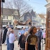 More parishioners are walking under the Epitaphios as they re-enter the church, at the end of the procession. Part of one of the wooden doors is visible in the upper right, and neighborhood houses and cars fill the background.