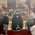 The four men and Fr. Aleksey are placing the Epitaphios back onto the tomb, which is located where the center table normally sits. The tomb is surrounded by flowers. Part of another person in a white shirt is visible in the lower right corner.