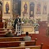Wearing a black vestment, Fr. Aleksey stands at the front left corner of the tomb, and he's facing the congregation. Some pews are seen in the lower left portion of the frame with two people seated. There's also a video camera on a tripod in front of Fr. Aleksey; the camera is live-streaming the service. There are flowers around the tomb, and a large wooden cross (about three feet tall?) at the back of the flowers.