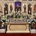 A close-up of the tomb, surrounded by flowers, with a large wooden cross behind the flowers. The iconostasis i picture in the background, and there are purple curtains across the archway of, and behind, the closed Royal Doors.