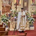 A medium shot of Fr. Aleksey standing in front of the tomb. His head is down as he looks at a bible in his hands, and he's facing the congregation. He's now wearing a white vestment. Some pews and a video camera on a tripod appear in the lower left, and the iconostas is in the background. Flowers surround the tomb, and there's a large wooden cross behind the flowers. This photo is much brighter than the others.