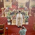 Another medium shot of Fr. Aleksey standing in front of the tomb, but this one was taken from the choir. He's facing the congregation and holding the Royal Gifts and he has his head down in prayer. He's wearing a white vestment, and a man in a green shirt is standing about five feet in front of him and is holding a candle. Some pews and a video camera on a tripod appear in the lower left, and a tall cross and part of a pew are along the right edge, with the iconostas is in the background. Flowers surround the tomb, and there's a large wooden cross behind the flowers. This photo is also very bright, and there are beams of light streaming from the chandelier at the top, about a third of the way in, and leading to the middle left of the frame.