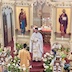 Fr. Aleksey is holding the Royal Gifts as he stands in front of the open Royal Doors. He's wearing a white vestment, and the Royal Gifts are covered by cloths that match his vestments. A young altar server in gold vestments is standing in front of Fr. Aleksey and facing him. A mural of the Virgin Mary and child appears to the left of the Royal Doors, and a mural of Jesus is on the right. There are potted flowers (white lilies, red tulips with orange tips, and purple flowers) placed on tables and around icons in the bottom third of the photo.
