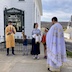 A closer-in version of the previous photo. Fr. Aleksey apperas towards the lower left of frame, and his back and left side are visible. Standing in front of him are, from left to right, an altar server holding a gold candelabra and cross, a young boy holding a wooden cross, and a teenage girl holding a white cloth and gold vessel of holy water. A younger girl holding an orange cloth is parially visible.