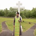 Fr. Aleksey stands outside at the church cemetary in front of a large, marble cross. He's facing the camera, and the cross towers several feet above him. He's wearing a green vestment with gold trim, and he's looking down at a prayer that he's holding. There's a, oval macadam walking path that goes around the cross and it's corresponding gravesite. There's grass both inside and outside the oval path, and a stone wall in that backgound that run perpendicularly across the middle of the frame, with trees on the far side of the wall. The sky is cloudy and appears very white in the photo.
