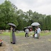 A view of the cemetary with Fr. Aleksey and several parishioners holding umbrellas as they pray. Fr. Aleksey is standing in the bottom-left corner, facing a gravesite that is out-of-frame, although a few flowers are visble. The parishioners are standing behind him in the lower right-hand quarter of the photo and looking down at papers in their hand as they pray. Grave sites, grass, and trees are visible in the background, along with a cloudy sky.