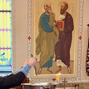 Photo of a wall mural of Sts. Peter and Paul. There's a large, silver bowl of holy water and three small, goldish candles in the foreground, and Fr. Aleksey's arms, covered by black cassock sleeves and light-blue cuffs, appear in the lower-left corner of the photo as he holds a cross in his hands above the bowl.