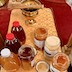 On a table with a gold, patterned cloth, there's a gold bowl with a holy water brush resting across the top, along with four open bottles of honey and three squeeze bottles of honey. Several different varieties and shades of honey are shown. There's a clear sheet of either plastic or glass on top of the gold cloth to protect it.