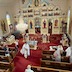 View from the choir loft showing Fr. Aleksey walking down the center aisle as he blesses members of the congregation who are standing within the pews.