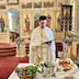 Fr. Aleksey is facing the camera and standing behind a table that appears in the lower quarter of the frame. The table is covered by a white cloth and has a metal collander and two baskets that all contain fruit and/or vegetables. He's looking at  a large, black prayer book that he's holding in his left hand. His right hand holds a holy water brush that's resting on the edge of a gold vessel that contains holy water.