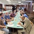 A series of long tables, covered by pastel-green tablecloths, running from bottom-center of the photo to roughly a third from the top. There are people seated on both sides of the table, and they're eating and talking. Part of another pastel-green table, running parallel to the first, is visible in the background on the left-hand side of the photo. The walls have dark paneling, and a chalkboard is against the back wall, at the end of the first table. One man is standing at the far end of the right-side of the table near the blackboard.