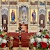 Fr. Aleksey is facing the altar while kneeling in the archway of the Royal Doors. He's wearing a red vestment with gold ornamentation and trim. Part of the iconostasis is also visible in the top two-thirds of the photo. There are flowers visible in both bottom corners of the photo — red roses and white carnations, with associated greenery. There is also a cross on the table, between the flowers, and vigil candles on either side of the cross.