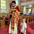 Fr. Aleksey is standing roughly in the center of the photo, facing left, and is seen blessing himself. There's a young boy standing to his left. He's holding a stuffed animal in his left hand and is looking at the camera. Some parishioners are seen looking onward as they stand in pews in the background.