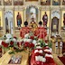 Fr. Aleksey is standing a few feet in front of the open Royal Doors holding the Royal Gifts and presenting them to the congregation. Two altar servers are standing in front of him, facing him, such that he appears between them. The center table, which holds crosses, an icon, candles, and flowers, is shown in the foreground.