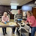 Three woman are working at a table and mobile cart, assembling paska breads. The woman on the left is making braids and twists out of dough pieces; the woman in the center is placing the braids and twists on the tops of loaves of paska bread that are in round, black baking pans; the woman on the right is holding a white cup in her left hand and a bright yellow brush in her right hand, and she's putting butter or oil on top of the completed dough loaves so they brown nicely in the oven. All three have paused their work and are looking at the camera and smiling.
