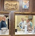A man and two women are standing in the hall kitchen, visible through two open windows between the main part of the hall and the kitchen, and they're smiling towards the camera. The man is shown through the left window, and there are two large, metal bowls on the counter between him and the window frame. There's a mixer, a measuring cup, and some kitchen tools on the counter between the women and the right window frame.